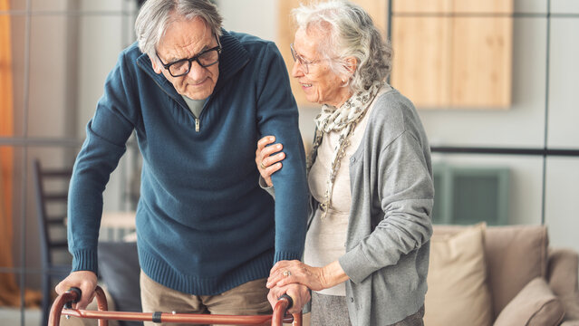 An elderly couple helps each other while using a walking frame