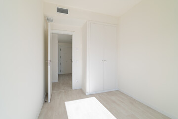 A bright, empty room with white walls and light wooden flooring. A closed door leads to another space, and a wardrobe is visible against the wall.