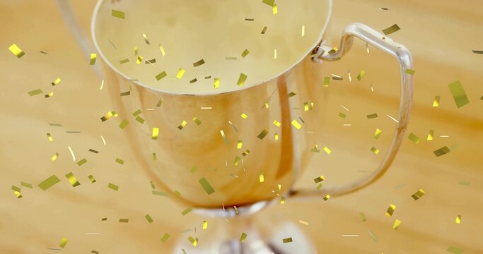 Shining silver trophy cup sitting on light wooden tabletop, gold confetti floating and resting