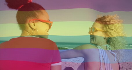 Talking women facing each other at beach, wearing shades, red tee, striped swimsuit, rainbow tint