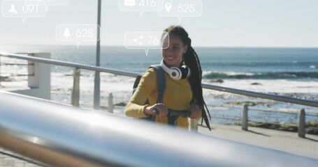 Standing woman wearing yellow top holding kick-scooter on coastal promenade, headphones and icons
