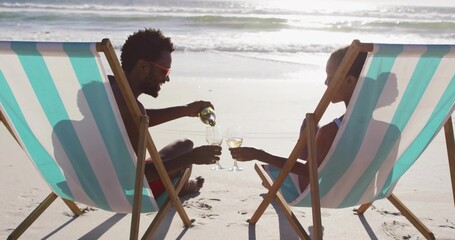 Naklejka premium Pouring couple in swimwear seated in striped deck chairs at shore, green bottle and wine glasses