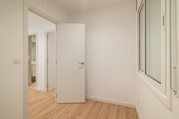 A modern, empty room with white walls and wooden flooring. A closed white door is visible, leading to another space. Natural light enters through a window.