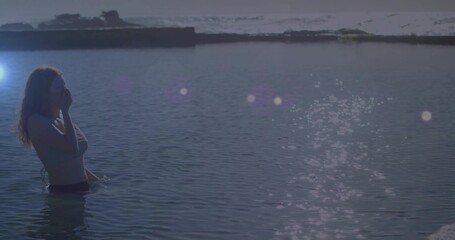 Standing woman wearing swimsuit touching face in shallow water near jetty, lens flare, copy space