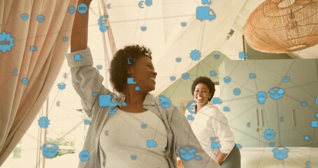 Smiling two mid adult Black women wearing white tops standing in bright bedroom, with blue icons