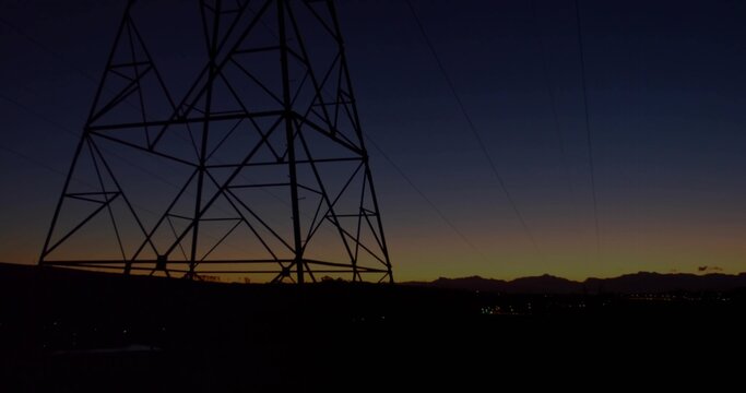 Dominating transmission tower looming left at dusk, sending power lines over ridge and town lights