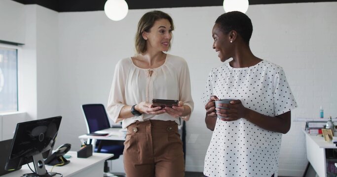 Discussing two coworkers wearing blouses, facing each other in modern office, holding phone and mug