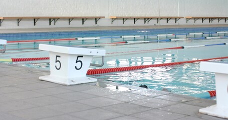 Showing white starting block numbered 5 sitting on tiled deck by calm pool, lane dividers visible