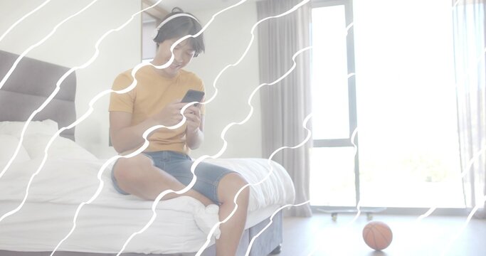 Sitting man in mustard tee, jean shorts, headset, using smartphone on bed by basketball, copy space