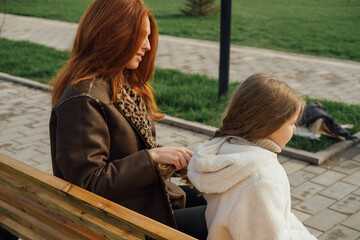 Mother braiding daughter hair on a park bench. Autumn setting, warm clothes, tender moment, outdoor.