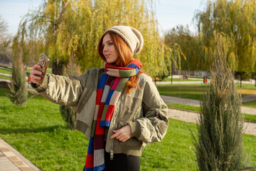 A young woman enjoying a sunny autumn or winter day outdoors, taking a cheerful selfie on a paved path in a park. Making photo.