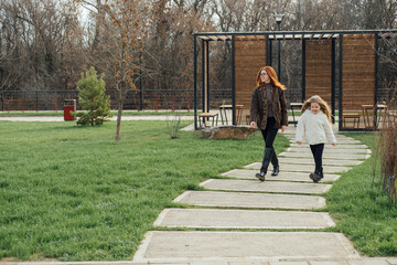Mother and daughter walking on wooden path through autumn park. Warm clothes, plush toy, peaceful moment.