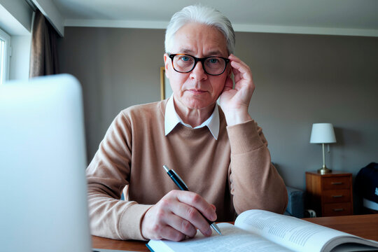 Portrait of senior Caucasian man wearing glasses sitting at desk studying open book and holding pen, looking directly into camera with thoughtful expression in home office setting - Powered by Adobe