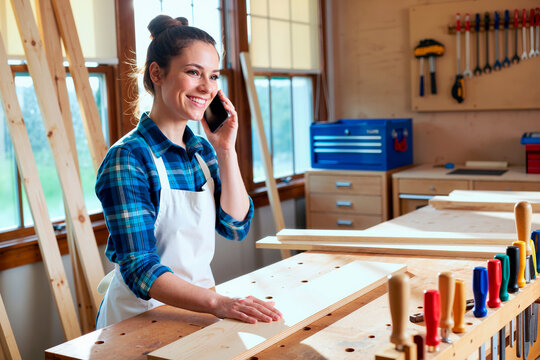 Caucasian young adult woman smiling while talking on smartphone and working with wooden boards in carpentry workshop, standing at workbench with tools visible in background