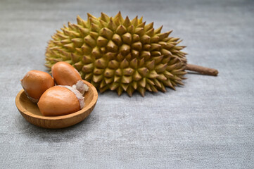 A spiky skin of durian rests in the background while three glossy durian seeds sit neatly in a small wooden bowl, highlighting the fruit’s rich texture and tropical character.