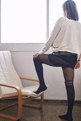 Woman in lingerie with her back to the camera, looking out of a window with one foot on a chair, vertical format.