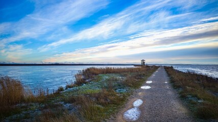 Scenic winter lakeside path with frozen water, dry reeds and bright sky leading to a distant wooden...