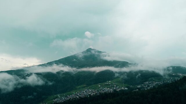 A cinematic aerial view of the majestic Mount Lawu, shrouded in mist and clouds at its peak with a small settlement on its slopes in Tawangmangu, Central Java, on Saturday, October 25, 2025.