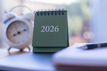 Close-up of green 2026 desk calendar with blurred clock and notebook in background, ideal for new year planning, time management, and productivity visuals.