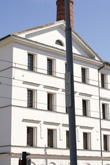 A bright white, historic building facade with neoclassical elements and dark rectangular windows, partially obscured by a tall street lamp post and a brick chimney on the roof