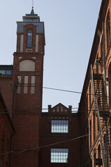 Obraz premium A tall, historic red brick factory tower with a contrasting light-colored section, framed by surrounding industrial buildings and a clear sky