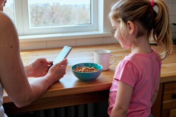 child girl standing at kitchen counter watching woman using smartphone near bowl of cereal and mug, illustrating morning routine and family care