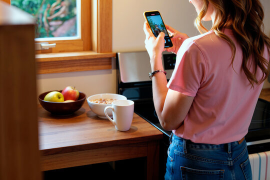 Young woman standing in kitchen using smartphone to photograph bowl of cereal and fruit on wooden counter, capturing healthy breakfast for medical care context