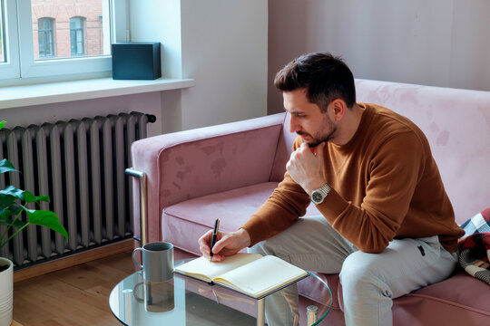 Young man sitting on sofa writing in notebook with thoughtful expression, holding pen in right hand, engaging in self reflection or planning for medical care - Powered by Adobe