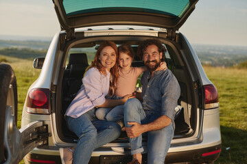 Family gathers around an open car trunk in a grassy area, smiling and enjoying the sunny day together. Laughter and warmth fill the atmosphere as they share this delightful time.