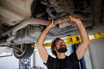 A skilled car mechanic works diligently under a raised vehicle in a busy auto repair shop. He inspects the exhaust system, demonstrating expertise and dedication to his craft.