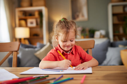 A young child is happily drawing on paper with colorful markers at a wooden table. The cozy living room is inviting, filled with soft lighting and family warmth.