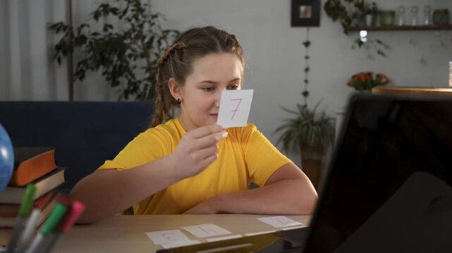 Girl studying with flashcard and laptop using number card recognition and learning at table smiling and arranging homework card set child engaged in education indoors with pencil and small plant