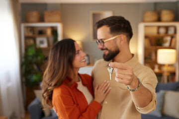 Joyful couple stands together in their new home, smiling as they hold up the keys. The comfortable living room features soft lighting and modern decor, creating a warm atmosphere.