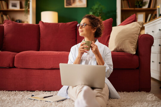 Person sitting on a floor with a laptop and mug, looking content in a cozy living room decorated with books and soft furnishings.