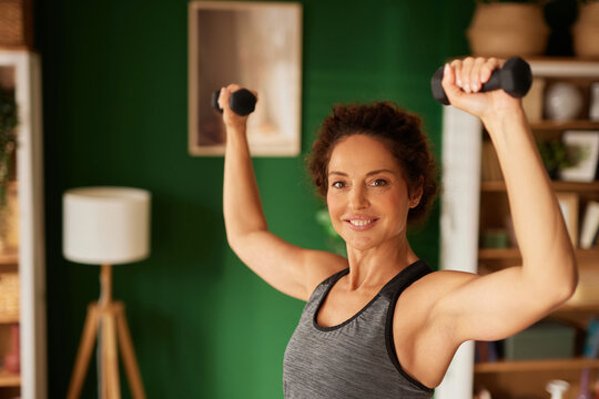 A woman lifts dumbbells above her head in a cozy home gym. The bright room features green walls and natural light, creating an inviting atmosphere for fitness.