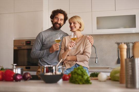 A couple smiles and holds glasses of wine in their modern kitchen, surrounded by fresh ingredients while preparing a meal together. They appear happy and relaxed in the warm atmosphere.