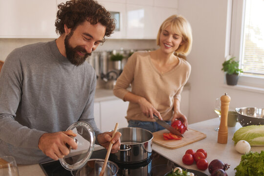 A man and woman are joyfully cooking in a contemporary kitchen. The man adds seasoning to a pot while the woman chops vegetables on a cutting board. Bright sunlight fills the space. - Powered by Adobe