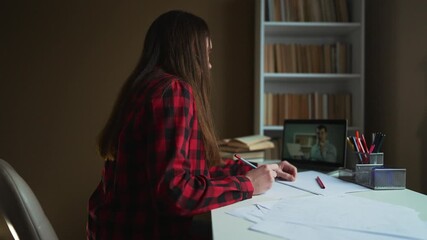 Studying online class with laptop teenager student sits at desk drawing on paper while following teacher on screen during homework session papers pencil holder bookshelf nearby focusing on study