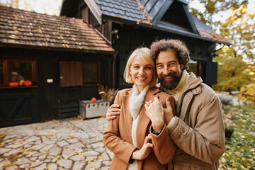 A couple stands together in a warm embrace, smiling happily during a weekend in autumn. Vibrant leaves fall around their cozy home, creating a perfect backdrop for family memories.