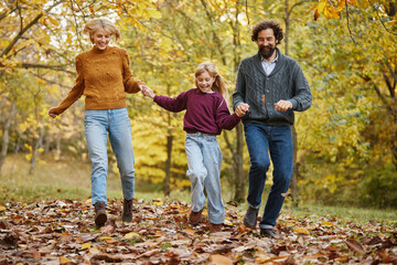 A family of three joyfully runs together in an autumn park, surrounded by vibrant yellow and orange leaves. They share laughter and playfulness on a warm weekend.