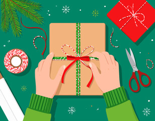 Top-down view of hands tying a red ribbon around a brown gift box, with wrapping supplies and Christmas decorations on a green surface.