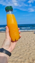 Hand holding a bottle of orange juice on a sunny beach with blue sea in the background. this image conveys refreshment and summer relaxation