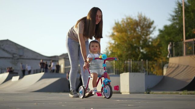 Woman guides toddler on tricycle near skatepark ramp with parent support and gentle smile while child practices balance and learning in outdoor urban area with tricycle wheel glinting under warm sun