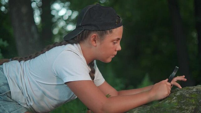 Girl kneels to study forest rock. Explorer child uses magnifier for research. Student repeats nature learning process. Magnifier helps child discovery. Forest becomes classroom explorer girl student