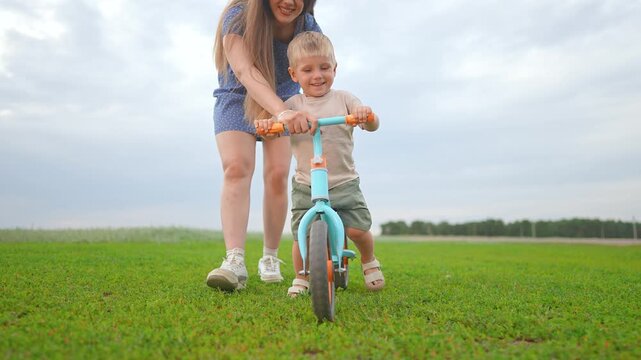 Mother walks beside boy teaching outdoor biking. Boy enjoys ride across field. Biking brings bonding time. Mother supports child learning to ride. Toddler stays focused during outdoor biking support