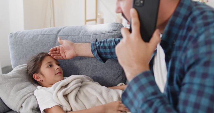 Father comforts daughter with fever at home. He checks the child, a girl, and calls a doctor by phone for advice on virus symptoms, illness and rest. Concept: father child telehealth at home.
