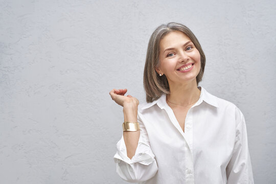 Smiling mature Caucasian female adult in white shirt posing indoors against light textured wall, raising hand with relaxed friendly gesture, expressing confidence and positivity - Powered by Adobe