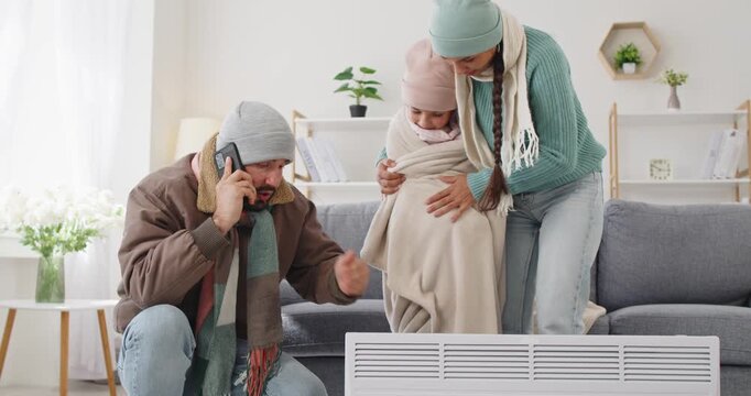 Family faces cold winter frost, seeking heating warmth. Parents help a child warm hands by a heater at home in room during a power outage noting safety and comfort. Warmth and safety at home.
