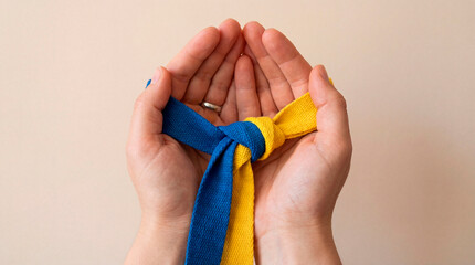 Close-up of two hands gently holding an intertwined blue and yellow ribbon on a clean neutral background. Symbol of unity, support, resilience and shared hope, expressing connection and solidarity.
