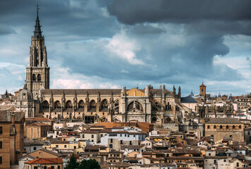 Aerial view of the imposing Toledo Cathedral piercing the skyline above a sea of terracotta rooftops under a dramatic sky, Toledo, Spain.